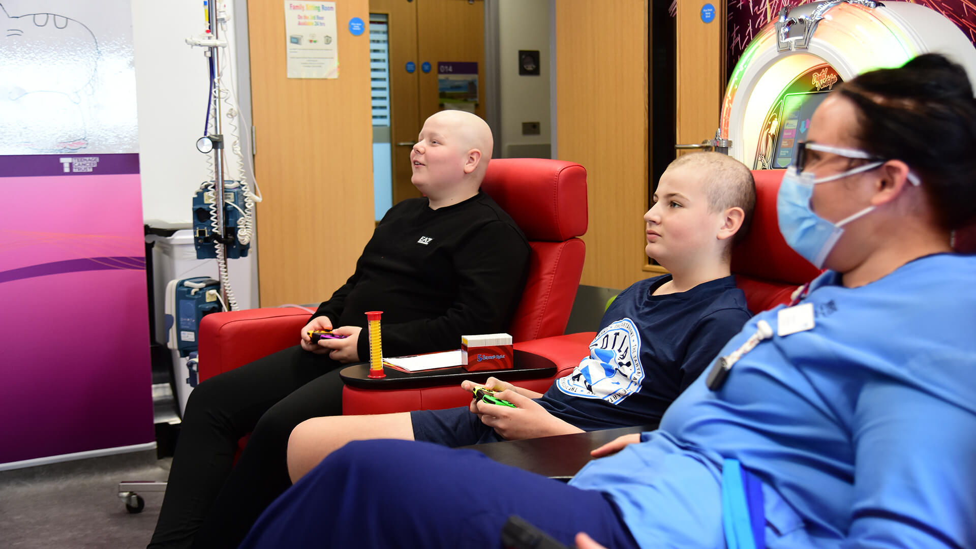 Two teenage boys playing video games while a Teenage Cancer Trust nurse watches on a Teenage Cancer Trust unit